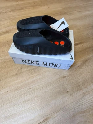 Svarta tofflor Nike Mind - Svarta tofflor från Nike Mind med vågig sula och ventilerande ovandel. Snygg och futuristisk design med små oranga detaljer på innersulan. Perfekta för chill dagar hemma eller när du vill ha något bekvämt på fötterna.