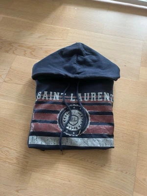 Mörkblå Saint Laurent hoodie - Snygg mörkblå hoodie från Saint Laurent med tryck framtill i vitt och rött, samt texten 'SAINT LAURENT' och ett emblem. Hoodien har huva med dragsko och en stor magficka. Perfekt för en avslappnad och trendig stil.
