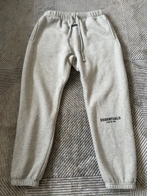 Grå Essentials Fear of God mjukisbyxor - Snygga grå mjukisbyxor från Essentials Fear of God med dragsko i midjan och resår vid bensluten. Byxorna har diskret logga på benet och är gjorda i mjukt sweatshirtmaterial. Perfekta för en avslappnad och trendig look.