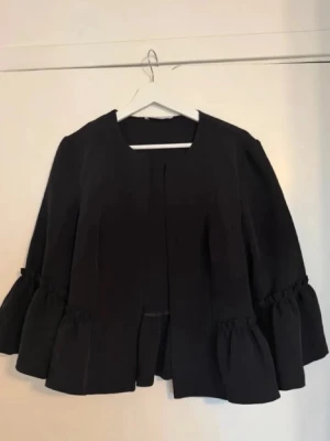 Svart kavaj med volangärmar - Säljer en svart kavaj med vida volangärmar och öppen framsida. Kavajen har en loose passform och är perfekt för dig som vill ha en unik twist på din outfit. Materialet känns mjukt och följsamt, och modellen är riktigt trendig. Så himla elegant!