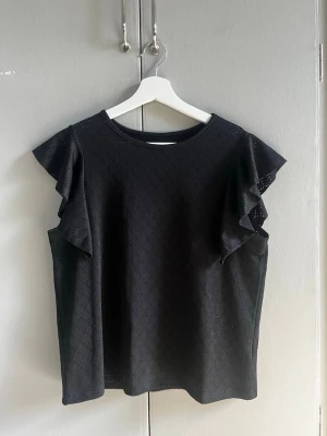 Svart blus med volangärmar - Snygg svart blus med volangdetaljer på ärmarna och diskret hålmönster i tyget. Blusen har rund halsringning och är tillverkad i ett lätt material som ger en luftig känsla. Perfekt för dig som vill ha en stilren topp med det lilla extra.