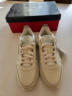 Vita Reebok Classic sneakers - Snygga vita Reebok Classic sneakers med chunky sula och diskret logga på plösen. Skorna har en låg silhuett, vita skosnören och ovandel i läder. Perfekta för dig som gillar retrostil och vill ha något stilrent till vardagslooken.