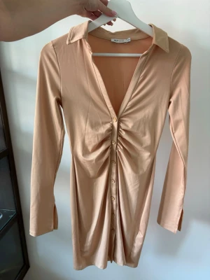 Beige klänning - Snygg beige kortklänning med lång ärm och v-ringning. Klänningen har knappar framtill och lätt rynkning vid bysten. Använd enstaka gånger. 