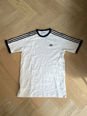 OANVÄND Beige Adidas t-shirt ”3S CALI T-SHIRT” med bruna detaljer - Klassisk beige t-shirt från Adidas med bruna ränder längs axlarna och bruna muddar vid ärmslut och hals. Adidas-logga på bröstet. Perfekt till streetwear och enkel att matcha med allt. Tillverkad i mjuk bomull för skön känsla. Modell 3S CALI T-SHIRT, säljs pga fel storlek.