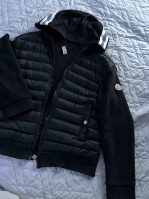 Svart Moncler cardiganjacka med huva - Moncler Cardigan perfekt nu inför hösten. Den är i mycket bra skick. Jackan köptes på monclers hemsida för ca 3 år sedan men har sedan dess använts få gånger. Har tyvärr inget kvitto men har en bild på scannen. 