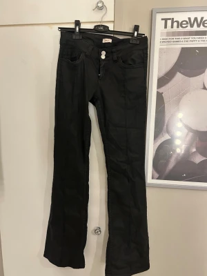Svarta bootcut jeans med dubbla knappar - Snygga svarta bootcut jeans med dubbla knappar i midjan och klassiska fickor både fram och bak. Jeansen har en lätt glansig finish och är perfekta för dig som gillar en stilren look med lite edge.