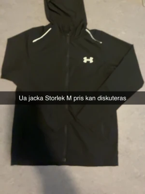 Svart Under Armour vindjacka med huva - Snygg svart vindjacka från Under Armour med huva och dragkedja framtill. Jackan har vita detaljer vid axlarna och logga på bröstet samt stor UNDER ARMOUR-text på ryggen. Perfekt för blåsiga dagar och sportiga outfits.