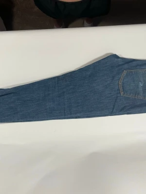 Blå jeans från Nudie Jeans - Säljer ett par klassiska blå jeans från Nudie Jeans med slim ben och snygga kontrastsömmar. Jeansen har två bakfickor med vågigt broderi och en läderpatch i midjan. Perfekta för dig som gillar en tidlös och avslappnad stil.
