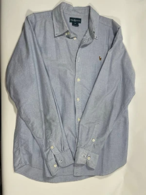 Ljusblå skjorta från Ralph Lauren - Klassisk ljusblå skjorta från Ralph Lauren med button-down krage och broderad logga på bröstet. Skjortan har långa ärmar, vita knappar och är tillverkad i mjuk bomull. Perfekt för en clean och stilren look.