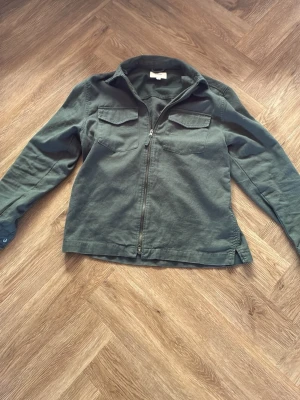 Mörkgrön overshirt från Dobber - Stilren mörkgrön overshirt från Dobber med dragkedja framtill och två bröstfickor med lock. Tillverkad i ett mjukt bomullstyg och har klassisk krage samt långa ärmar med knapp vid ärmslut. Perfekt lager på lager-plagg för en avslappnad look. Använd 3 gånger, nypris 999