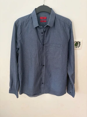Blå skjorta med små prickar från Hugo boss - Stilren blå skjorta med små vita prickar och klassisk krage. Skjortan har lång ärm och knäppning framtill. Perfekt för dig som gillar en enkel men ändå snygg look.