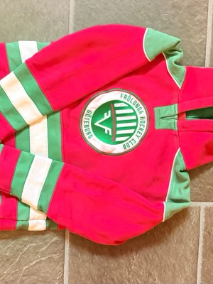 Frölunda HC hoodie röd och grön - Snygg hoodie i rött, grönt och vitt med Frölunda Hockey Club Göteborg-logga på bröstet. Tröjan har huva och breda ränder på ärmar och nederkant. Perfekt för dig som vill visa ditt stöd för Frölunda och sticka ut i mängden.