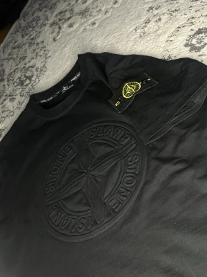  Stone Island  - Snygg svart t-shirt från Stone Island med stor ton-i-ton broderad logga på bröstet och den klassiska patchen på ärmen. T-shirten har rund hals och är tillverkad i mjuk bomull. Perfekt för dig som gillar streetwear och vill ha något stilrent men ändå med edge.