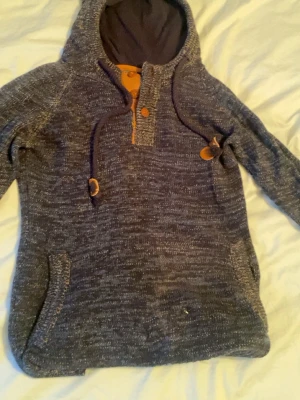 Mörkgrå hoodie med knappar och huva - Mysig mörkgrå hoodie med melerad look, stor huva och knappar framtill. Dragsko med trädetaljer och fickor på sidorna. Perfekt för en avslappnad stil och passar till jeans eller mjukisbyxor.