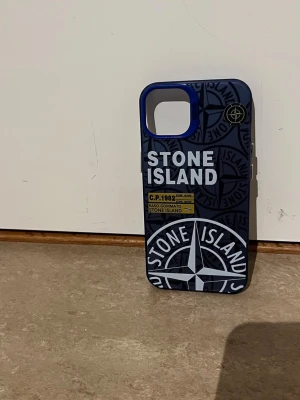 Blått Stone Island mobilskal - Snyggt mobilskal i blått med Stone Island-logga och grafiskt mönster. Skalet har en tydlig logotyp och detaljer i vitt och gult, samt en blå ram runt kamerahålet. Tillverkat i hårdplast och passar perfekt för dig som vill sticka ut.
