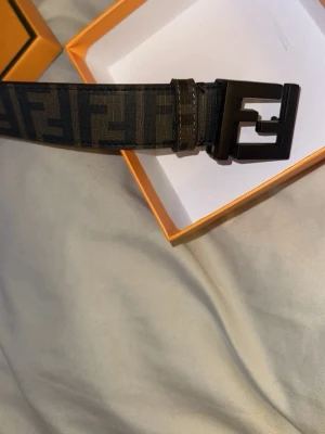 Svart och brun Fendi bälte med logga - Snyggt bälte från Fendi i svart och brun med det klassiska FF-logomönstret. Bältet har ett stort svart spänne med Fendi-logga och levereras i original ask. Perfekt accessoar för att lyfta din outfit.
