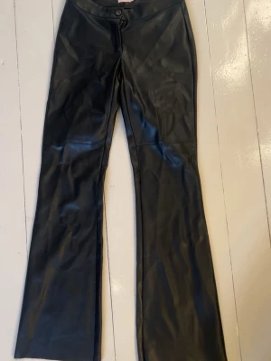 Svarta utsvängda byxor i fuskskinn - Säljer ett par svarta byxor i fuskskinn med utsvängda ben och mid waist. Byxorna har sömdetaljer över knäna och stängs med snörning framtill. Perfekta för en edgy och trendig look. Sitter riktigt snyggt på, skriv vid bild där byxorna sitter på. Väljer att sälja dessa för att de inte riktigt är min stil och fick dem i present så har testat några gånger hemma men aldrig använts till vardags.