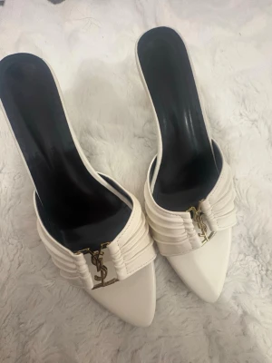 Vita spetsiga pumps med gulddetalj - Säljer ett par vita pumps med spetsig tå och smal klack. Skorna har en snygg guldfärgad logga YSL framtill och veckade detaljer på ovandelen. Insidan är svart och materialet ser ut att vara slätt läder eller konstläder.