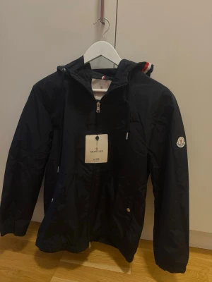 Moncler windbreaker - Snygg svart vindjacka från Moncler med huva och dragkedja framtill. Jackan har Moncler-logga på ärmen och detaljer i rött, vitt och blått på huvan. Tillverkad i lätt och tåligt material, perfekt för blåsiga dagar.