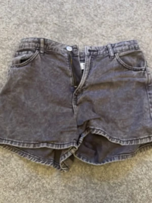 Grå denimshorts med hög midja - Säljer ett par grå denimshorts med hög midja och klassisk femficksdesign. Shortsen har knapp- och dragkedjestängning samt råa kanter nedtill för en avslappnad look. Perfekta till sommaren och lätta att matcha med olika stilar.