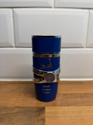 Asad Zanzibar parfym blå flaska - Snygg parfymflaska från Lattafa i en djupblå färg med silvriga detaljer och orientaliskt mönster. Flaskan har en modern, rundad form och text i guld. Perfekt för dig som gillar unika och stilrena dofter.