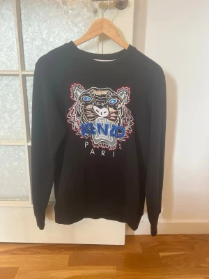 Svart stickad Kenzo-tröja med tiger - Svart stickad tröja från Kenzo med ikoniskt tigerbroderi i flera färger och texten 'KENZO PARIS' framtill. Tröjan har rund halsringning och långa ärmar. Perfekt för dig som vill sticka ut med en trendig och cool look.