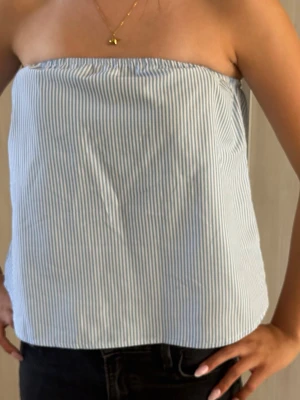 Blåvit randig bandeau tubtopp - Säljer en blåvit randig bandeau tubtopp med smala vertikala ränder. Toppen är axelbandslös och har en enkel, rak passform. Perfekt för varma dagar och enkel att matcha med jeans eller kjol. Sitter lite konstigt på modellen för att hon har bh under som banden är nedvikta på men annars sitter den helt underbart!