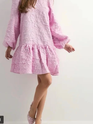 Ljusrosa kort klänning med snörning - Supersöt ljusrosa kort klänning med volangkant och ballongärmar. Klänningen har en öppen rygg med snörningsdetaljer och är gjord i ett strukturerat tyg som ger en unik look. Perfekt för dig som vill sticka ut med en feminin och trendig stil.