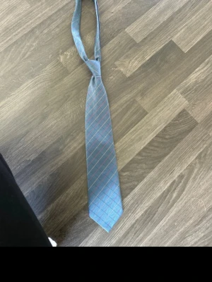 Blå slips med rutigt mönster - Snygg blå slips med tunna diagonala ränder i gult och rött som bildar ett rutigt mönster. Slipsen har klassisk form och är tillverkad i ett glansigt tyg som ger en stilren look. Perfekt för att lyfta vilken outfit som helst.