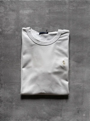 Vit t-shirt från Polo Ralph Lauren - Klassisk vit t-shirt från Polo Ralph Lauren med korta ärmar och rund hals. Diskret broderad logga i guld på bröstet. Tillverkad i mjuk bomull för en skön och avslappnad stil.