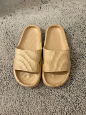 Beige ribbade slides sandaler - Säljer ett par beige slides sandaler med bred ribbad rem över foten. Tillverkade i ett mjukt och flexibelt syntetmaterial som ger skön komfort. Perfekta för chill dagar eller att glida runt hemma. Klassisk öppen tå och enkel design.