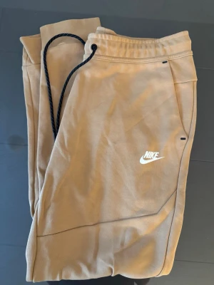 Beige mjukisbyxor från Nike - Snygga beige mjukisbyxor från Nike med svart dragsko i midjan och vita Nike-loggan på benet. Byxorna har avslappnad passform, resår i midjan och sidofickor. Perfekta för chill dagar eller när du vill ha en sportig look.