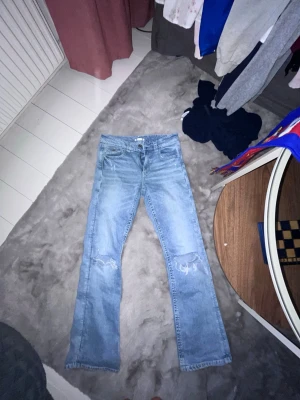 Ljusblå slitna jeans från Gina Tricot - Ljusblå jeans från Gina Tricot i rak modell med slitningar över knäna. Klassisk femficksdesign och knappgylf. Perfekta för en avslappnad och trendig look. Tillverkade i denim med snygg tvättad finish.