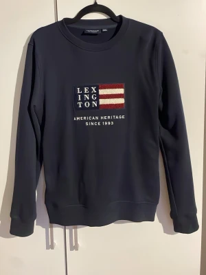 Mörkblå sweatshirt från Lexington - Mörkblå sweatshirt från Lexington med broderad amerikansk flagga och texten 'American Heritage Since 1993' på bröstet. Tröjan har rund halsringning, långa ärmar och ribbade muddar. Perfekt för en avslappnad och stilren look. Mycket bra kvalitet i 100% bommul