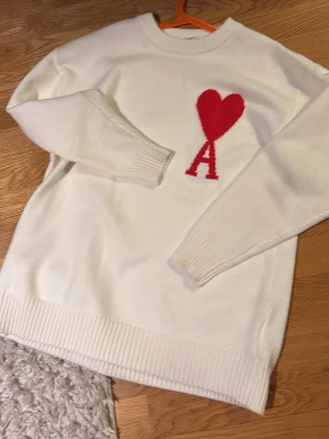 Vit stickad tröja med rött hjärta - Vit stickad tröja med rund hals och ribbade muddar. På bröstet finns ett rött hjärta och bokstaven A i rött. Tröjan har en avslappnad passform och är perfekt för dig som gillar enkel men snygg design.