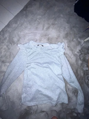 Ljusgrå tröja med volang från ONLY - Supersöt ljusgrå tröja från ONLY med volangdetaljer på axlarna. Tröjan har rund halsringning och långa ärmar. Perfekt för dig som gillar en enkel men ändå trendig look. Materialet känns mjukt och bekvämt mot huden.