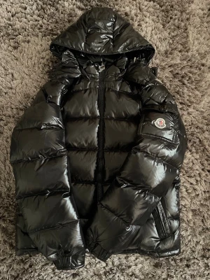 Svart glansig dunjacka med huva - Snygg svart dunjacka med glansig finish och huva. Jackan har långa ärmar, quiltad design och dragkedja framtill. Moncler-logga på ärmen ger en exklusiv touch. Perfekt för kalla dagar när du vill hålla stilen.