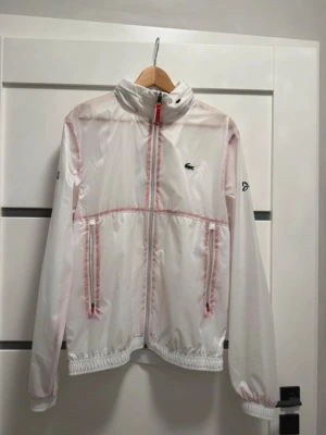 Lacoste vindjacka  - Snygg vit Lacoste x djokovic vindjacka med röda dragkedjor och detaljer. Jackan har hög krage, två fickor med dragkedja och elastiska muddar vid ärmslut och midja. Liten logotyp på bröstet och diskreta sömmar i rött. Perfekt för en sportig och trendig look. Storlek S/M LUVA KOMMER MED