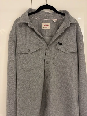 Grå overshirt från Wrangler - Snygg grå skjorta från Wrangler med diskret fiskbensmönster. Skjortan har två bröstfickor med knappar och klassisk krage. Perfekt för en avslappnad men ändå stilren look.