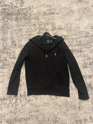 Svart hoodie från Polo Ralph Lauren - Snygg svart hoodie från Polo Ralph Lauren med dragkedja framtill och två fickor. Klassisk liten logga broderad på bröstet. Tillverkad i mjukt material och har justerbar huva med snören. Perfekt för en avslappnad och stilren look.