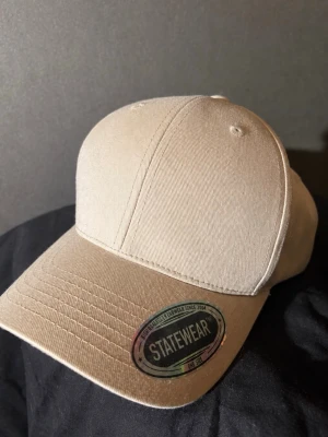 Beige keps från Statewear - Snygg beige keps från Statewear med klassisk böjd skärm och justerbar baksida. Tillverkad i slitstarkt bomullstyg och har en enkel, stilren design utan tryck eller brodyr. Perfekt för att komplettera din streetwear-look.