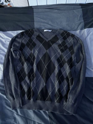 Grå argyle stickad tröja Dressmann - Mörkgrå stickad tröja från Dressmann i storlek M. Tröjan har V-ringning och klassiskt argylemönster i svart och grått framtill. Ribbstickade muddar vid ärmslut och nederkant. Perfekt för en stilren och tidlös look.