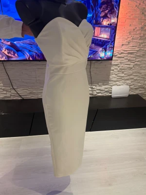 Vit axelbandslös midiklänning - Säljer en elegant vit axelbandslös midiklänning med figurnära passform och draperad detalj vid bysten. Klänningen har en stilren design och slutar under knät. Perfekt för dig som vill ha en clean och chic look.
