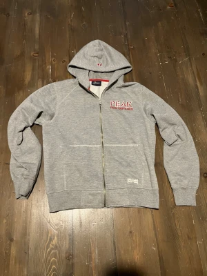 Grå vintage zip-hoodie Peak Performance - Snygg grå vintage zip-hoodie från Peak Performance med röd och vit logga på bröstet och broderad detalj på huvan. Mjuk insida, två fickor fram och ribbade muddar. Perfekt för chill dagar eller till träningen. Storlek L men sitter som M.