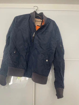 Alpha industries bomberjacka - Snygg mörkblå bomberjacka med klassiskt orange foder. Jackan har ribbade muddar vid ärmslut, krage och nederkant samt fickor med tryckknappar och en ärmficka med dragkedja. 