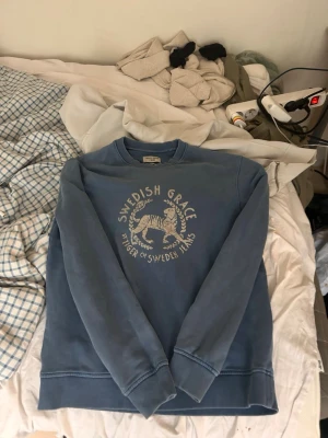 Blå sweatshirt från Tiger of Sweden - Blå sweatshirt från Tiger of Sweden Jeans med trycket 'Swedish Grace' och en tiger på bröstet. Tröjan har rund hals, ribbade muddar vid ärmslut och nederkant samt en avslappnad passform. Perfekt för en chill och stilren look.