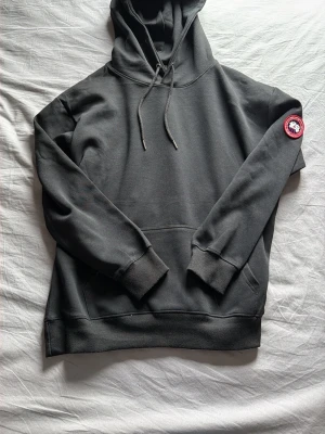 Svart hoodie från Canada Goose - Snygg svart hoodie från Canada Goose med klassisk huva och dragsko. Har en stor känguruficka framtill och den ikoniska Canada Goose-loggan på vänster ärm. Perfekt för en chill och stilren look.