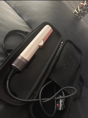 Dyson airstraight - Säljer en snygg plattång i rosa och vitt med digital display och flera inställningsknappar. Kommer i ett praktiskt svart fodral med dragkedja och nätficka för sladd och tillbehör. Perfekt för att styla håret snabbt och enkelt.