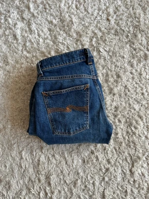 Blå raka jeans med kontrastsöm - Säljer ett par klassiska Nudie jeans i modellen solid ollie. Som är en modell med relativt raka ben och snygga kontrastsömmar på bakfickan. Jeansen är använda en gång och har inga defekter. Hör av er vid minsta lilla fråga