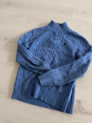 Blå kabelstickad tröja med halvzip Ralph lauren  - Kabelstickad blå tröja med halvzip och hög krage. Tröjan har långa ärmar, ribbstickade muddar och en liten broderad logga på bröstet. Perfekt för lager-på-lager och en snygg vardagslook.